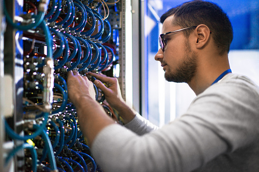 Side  view portrait of young man connecting cables in server cabinet while working with supercomputer in data center